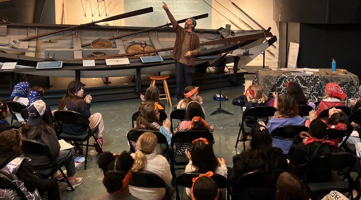 Audience in chairs watches a speaker standing in front of a boat in museum.