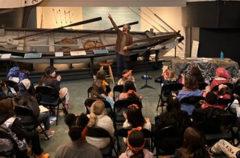 Audience in chairs watches a speaker standing in front of a boat in museum.